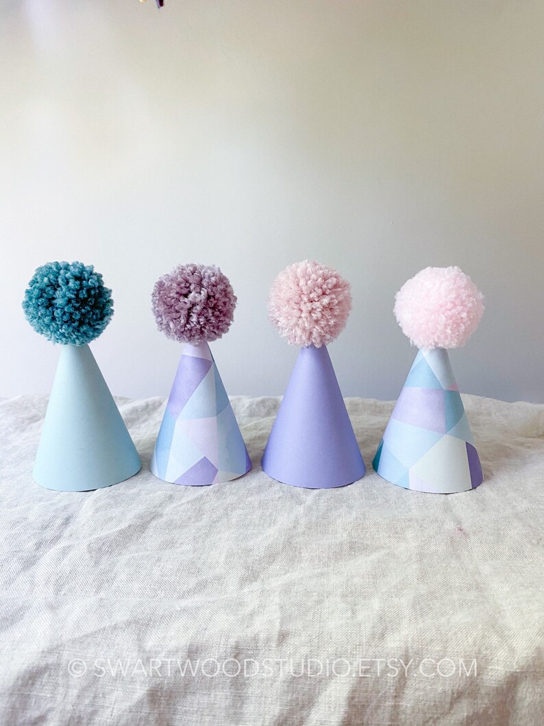 May include: Four party hats with pom poms on top. The hats are blue, purple, lavender, and pink. The hats have a geometric pattern.