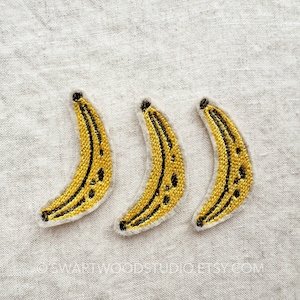May include: Three embroidered banana patches. Each patch features a yellow banana with black details and a white border. The patches are arranged in a row on a light gray fabric background. The text "SWARTWOODSTUDIO.ETSY.COM" is visible.