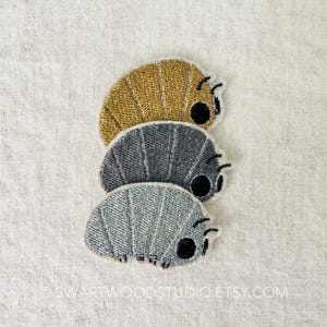 May include: Three embroidered pill bug patches in gold, gray, and silver, stacked on top of each other. Each bug has a black eye and small legs. The patches are on a white, textured background.