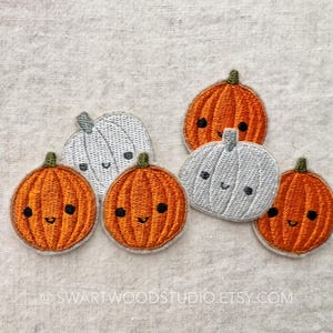 May include: Embroidered pumpkin patches in orange and gray with smiling faces and green stems. The patches are arranged on a neutral fabric background, perfect for fall-themed crafts and decorations.