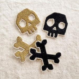 May include: Close-up of four embroidered patches: two skulls and two crossbones. One skull and one crossbones are gold with white trim, the other skull and crossbones are black with white trim. The patches are on a neutral linen background.