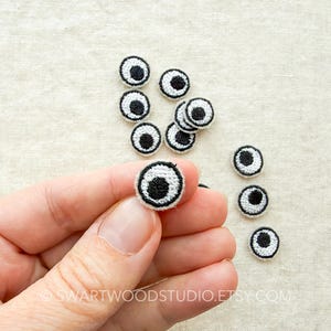 May include: A collection of small, circular embroidered patches featuring a white sclera, a black iris, and a black pupil, designed to resemble googly eyes. The patches have a stitched border and are arranged in a group with one held in the foreground.