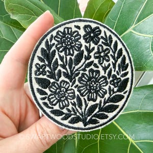 May include: A circular embroidered patch featuring a black floral and leaf pattern on a cream-colored background. The design includes various stylized flowers and foliage arranged in a dense, symmetrical composition with a thick black stitched border.
