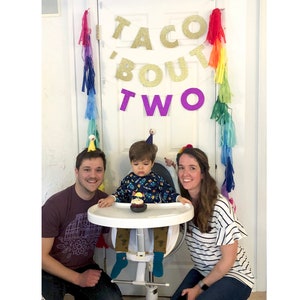 May include: A family celebrates a child's second birthday with a "Taco 'Bout Two" banner, rainbow streamers, and a birthday hat. The child sits in a high chair with a cupcake.