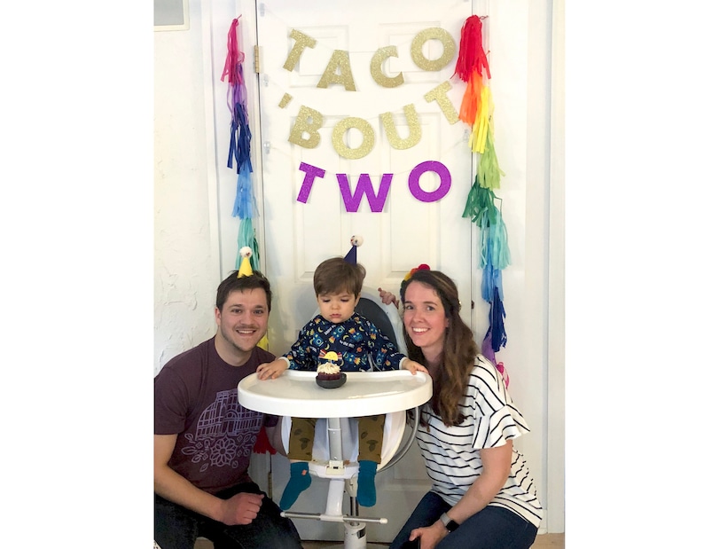May include: A family celebrates a child's second birthday with a "Taco 'Bout Two" banner, rainbow streamers, and a birthday hat. The child sits in a high chair with a cupcake.