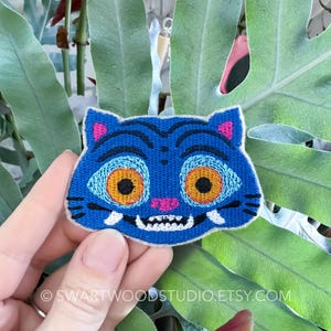 May include: Embroidered patch in the shape of a blue cat's face with orange eyes, pink ears, and white teeth. The patch is held in front of large green leaves. The patch has a stitched design and is likely for clothing or accessories.