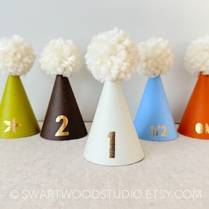 May include: Five colorful party hats with white pom-poms on top. The hats are decorated with gold numbers and a star. The numbers on the hats are 1/2, 1, 2, and "ONE".