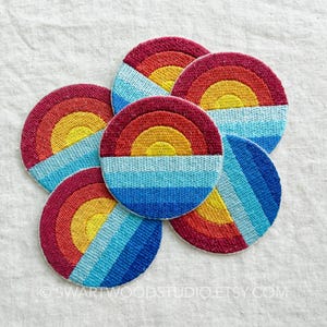 May include: Five circular embroidered patches featuring a stylized sun rising over a horizon. Each patch displays a yellow sun center, a red arch, and horizontal bands of light blue and dark blue thread. The text swartwoodstudio.etsy.com is visible.