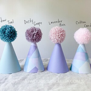 May include: Four party hats with pom-poms on top. The hats are decorated with a geometric pattern. The pom-poms are in the colors: Meadow Teal, Dusty Grape, Lavender Rose, and Cotton Candy.