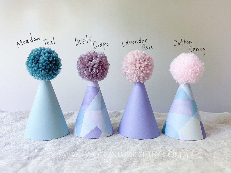 May include: Four party hats with pom-poms on top. The hats are decorated with a geometric pattern. The pom-poms are in the colors: Meadow Teal, Dusty Grape, Lavender Rose, and Cotton Candy.