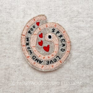 May include: A pink felt worm with red hearts and the text "Spread Love and Worm Poo" embroidered on it.