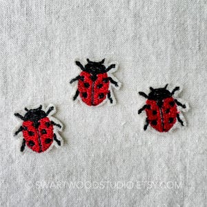 May include: Three red and black embroidered ladybug patches on a light gray fabric background.