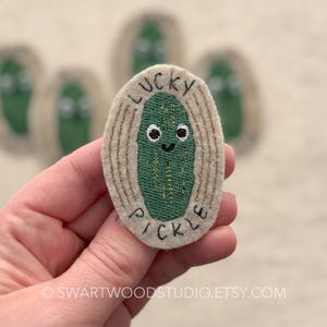 May include: A beige felt patch with gold embroidery. The patch features a green pickle with a smiling face and the text "LUCKY PICKLE".