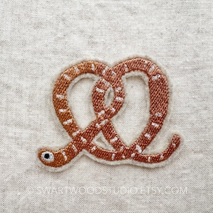 May include: A brown embroidered patch shaped like a pretzel. The patch is on a white linen background.