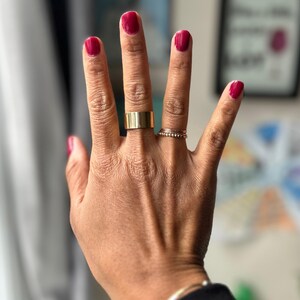 Può includere: Una mano con unghie smaltate e anelli d'oro. Le unghie sono dipinte di un rosso intenso. Un anello largo e piatto in oro è sul dito medio e un sottile anello di perline è sull'anulare. Un braccialetto d'argento è visibile sul polso.