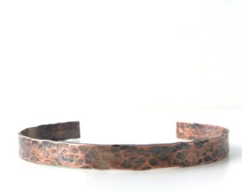 Puede incluir: Una pulsera de cobre tipo brazalete con una superficie martillada y texturizada con una pátina oscura y oxidada. La banda de metal tiene un diseño abierto, mostrando un acabado metálico marrón rojizo con hendiduras irregulares.