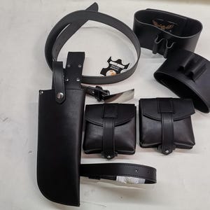 May include: A set of black leather accessories including a belt, a holster, and several pouches. The pouches have a flap closure and are designed to be worn on a belt.
