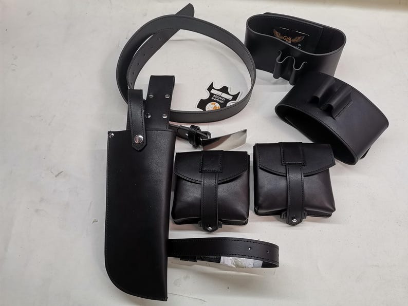 May include: A set of black leather accessories including a belt, a holster, and several pouches. The pouches have a flap closure and are designed to be worn on a belt.
