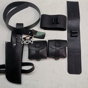 May include: A black leather belt with multiple pouches and a holster. The belt has a buckle and is decorated with a small leather tag that says "Genuine Leather".