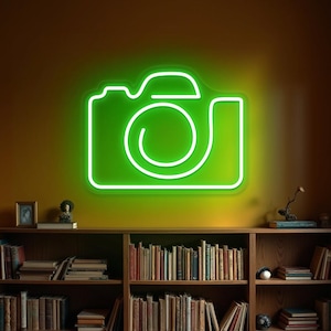Może przedstawiać: Neonowy, zielony znak w kształcie aparatu fotograficznego oświetla ścianę. Obrys znaku jest biały, z obiektywem i wizjerem. Poniżej znajduje się drewniany regał wypełniony książkami, tworzący ciepłą atmosferę. Znak jest elementem dekoracyjnym.