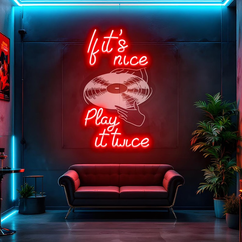 May include: A neon sign with the words "If it's nice, play it twice" in red script, above a white graphic of a vinyl record. The sign is mounted on a dark wall above a black sofa with gold legs. The room has a retro vibe.