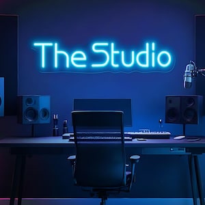 May include: A neon sign that reads "The Studio" in bright blue illuminates a recording studio. The studio features a desk with a computer, keyboard, and chair, along with speakers and a microphone. The background is a dark blue.