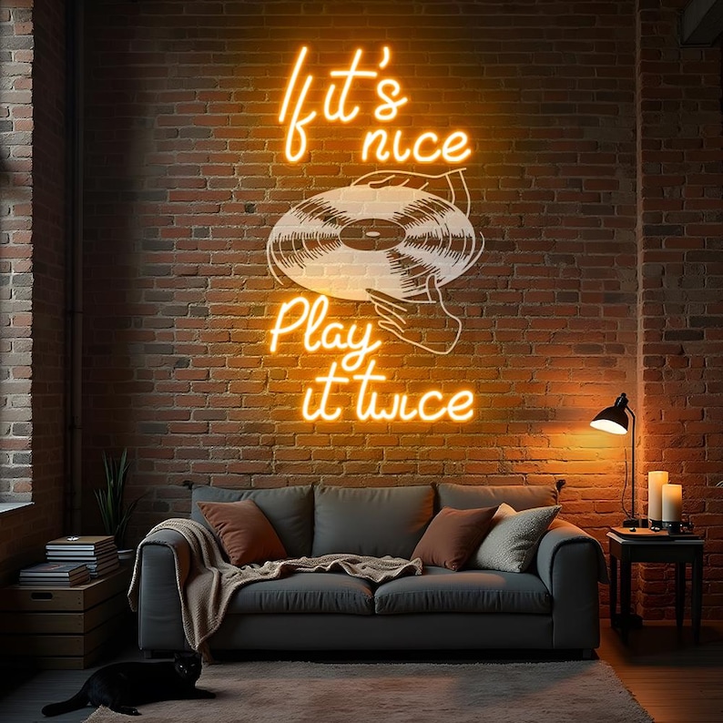 May include: Neon sign on a brick wall with the text "If it's nice, play it twice" and an illustration of a vinyl record. A gray sofa with throw pillows and a black cat are in the foreground. A small table with a lamp and candles is to the right.