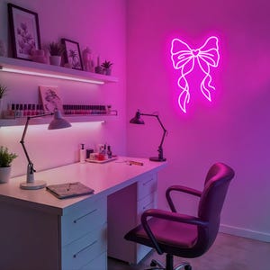 May include: A white desk with drawers and a black office chair in a room with pink walls. A pink neon sign of a bow with a heart in the center is on the wall.