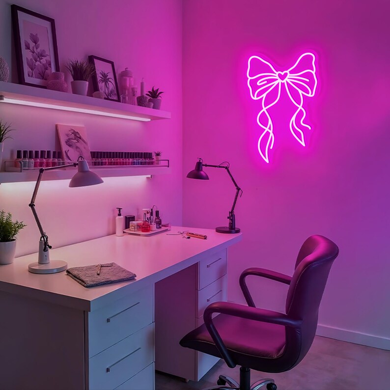 May include: A white desk with drawers and a black office chair in a room with pink walls. A pink neon sign of a bow with a heart in the center is on the wall.