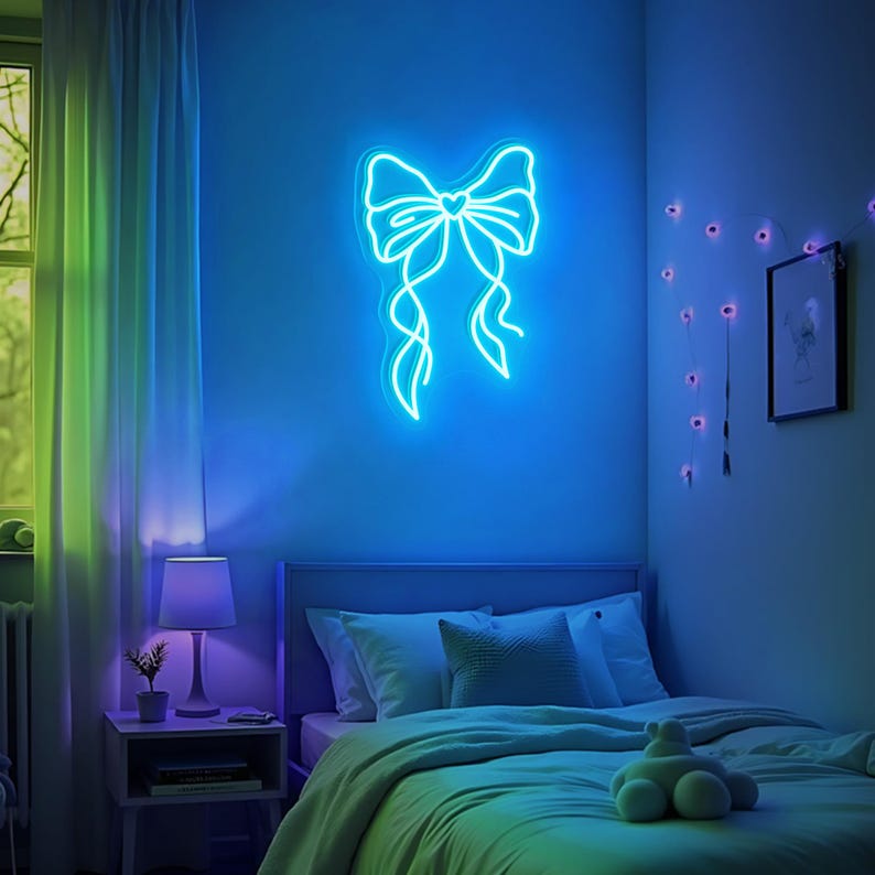 May include: A blue neon light shaped like a bow with a small heart in the center. The light is mounted on a wall in a bedroom with a bed, nightstand, and lamp.