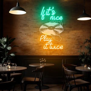 May include: A neon sign with the words "It's nice, Play it twice" in green and yellow, above a vinyl record illustration. The sign is mounted on a brick wall in a restaurant setting, with tables and chairs visible.