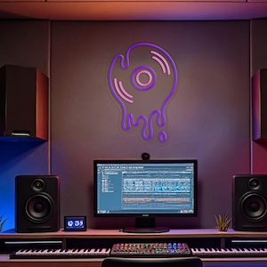 May include: A neon sign shaped like a melting vinyl record, with a purple outline and pink interior, hangs on a gray wall. The sign is above a desk with a computer monitor, speakers, and a keyboard, suggesting a music studio setup.