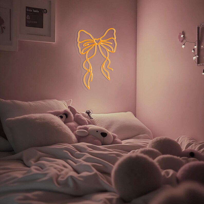 May include: A pink bedroom with a white bed and a neon yellow bow sign on the wall. The bed has white bedding and several fluffy pink pillows. There are also several stuffed animals on the bed.