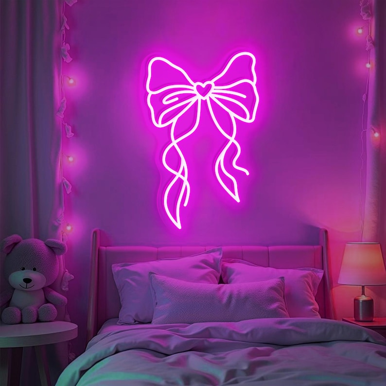 May include: A white neon sign shaped like a bow with a small heart in the center. The sign is lit up in a bright pink color and is mounted on a purple wall. The sign is in a bedroom setting with a bed, a nightstand, and a lamp.