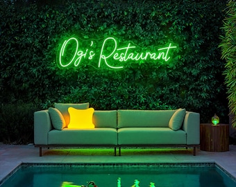 May include: A neon sign that reads "Ogi's Restaurant" in a cursive font is mounted on a wall of green foliage. Below the sign is a green sofa with yellow and green pillows. A pool with a duck float is in front of the sofa.