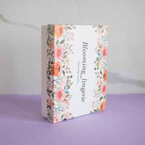 Puede incluir: Caja blanca con un diseño floral y el texto "Blooming_lingerie" y "@blooming_lingerie_" impreso en el lateral.