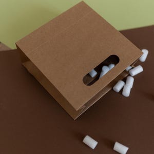 May include: A brown paper bag with a cut-out handle. The bag is lying on a brown surface and white packing peanuts are spilling out of the bag.