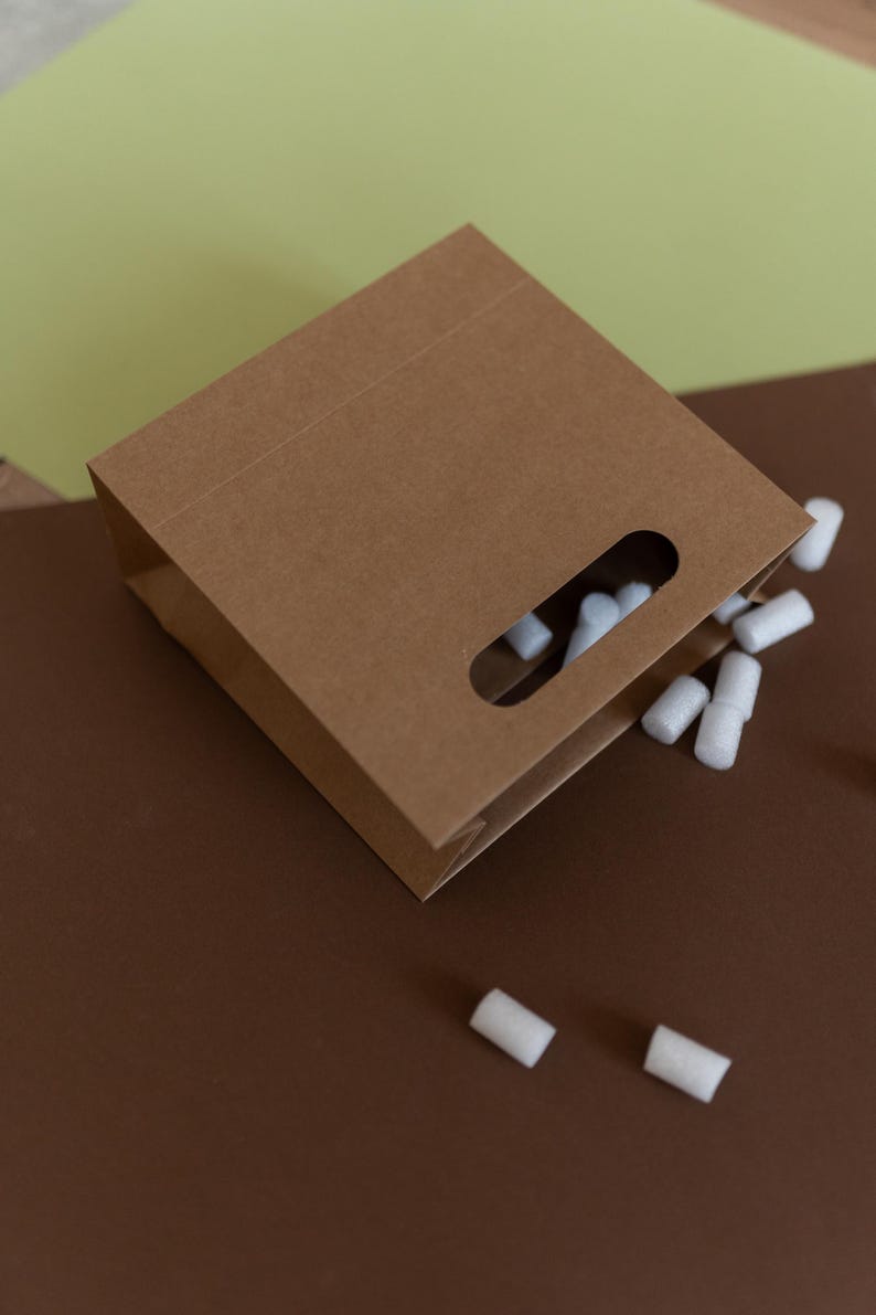 May include: A brown paper bag with a cut-out handle. The bag is lying on a brown surface and white packing peanuts are spilling out of the bag.