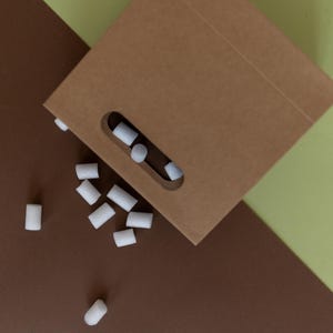May include: A brown paper bag with a cut-out handle is open and spilling out white foam packing peanuts onto a brown surface.