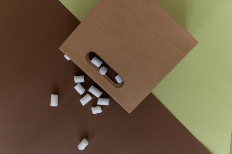 May include: A brown paper bag with a cut-out handle is open and spilling out white foam packing peanuts onto a brown surface.