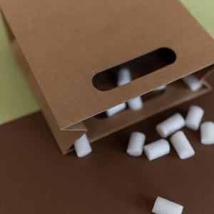 May include: A brown paper bag with a cut-out handle is spilling out white packing peanuts onto a brown surface.