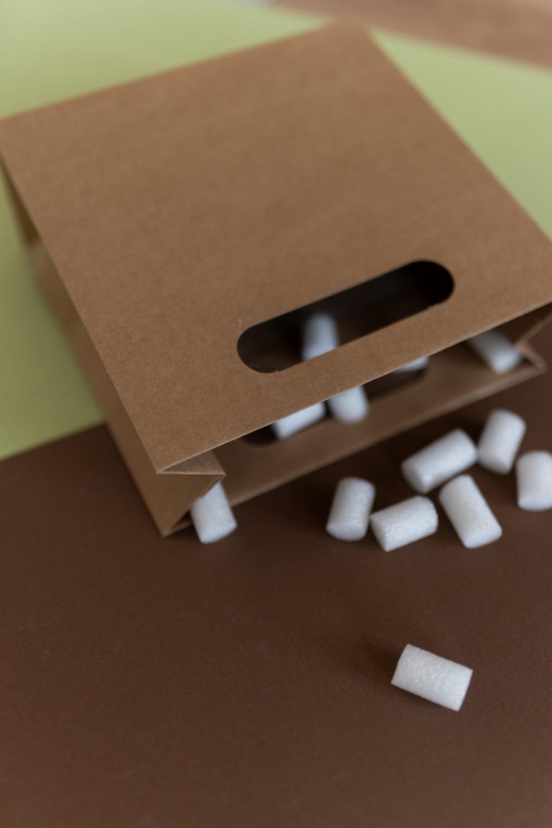 May include: A brown paper bag with a cut-out handle is spilling out white packing peanuts onto a brown surface.