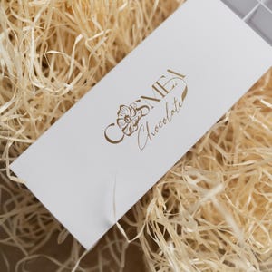 May include: A rectangular white cardboard box with a sliding tray featuring a gold foil logo that reads COSMEA Chocolate with a floral illustration. The box has internal dividers and is positioned at an angle on a bed of light tan wood shavings.