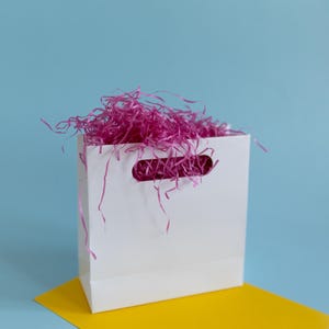 May include: A white paper gift bag filled with pink tissue paper. The bag is sitting on a yellow surface against a light blue background.