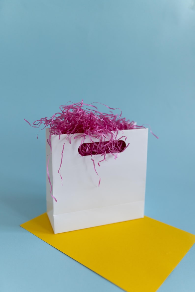 May include: A white paper gift bag filled with pink tissue paper. The bag is sitting on a yellow surface against a light blue background.