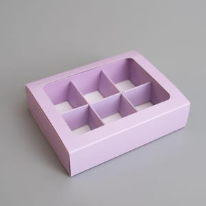 Peut inclure: Une boîte en carton de couleur lavande avec une fenêtre transparente, divisée en neuf compartiments. La boîte est conçue pour emballer de petits objets, tels que des chocolats ou des bijoux.