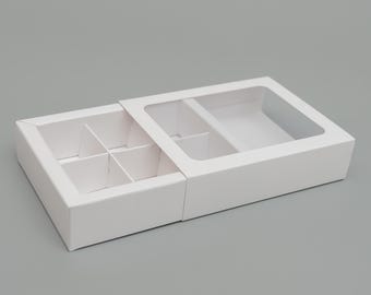 May include: White cardboard box with a clear plastic window, divided into six compartments. The box is open, revealing the compartments.