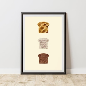 Op de afbeelding: Ingelijste kunstdruk met drie sneetjes brood: een goudbruine, een lichtgekleurde snee met de tekst "MAN SHALL NOT LIVE BY BREAD ALONE" en een donkerbruine snee. Het kunstwerk staat tegen een crèmekleurige achtergrond.
