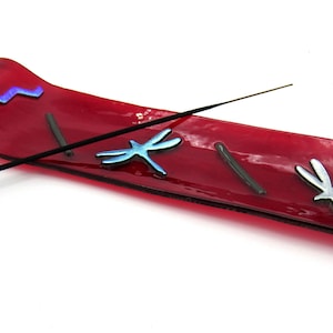 May include: A red glass incense holder with a wavy design and dragonfly accents. The holder features a zig-zag pattern in blue and silver dragonfly shapes. A black incense stick rests on the holder.
