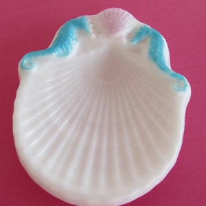 May include: A white seashell-shaped dish with a ribbed interior. The rim is accented with light blue and pink paint. The dish is likely made of ceramic or porcelain and is suitable for holding small items.